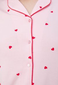 Rosa pyjamasöverdel med röda hjärt-mönster, knappknäppning och kontrasterande röd piping längs kragen och knappslån. Mjuk tygkänsla.
