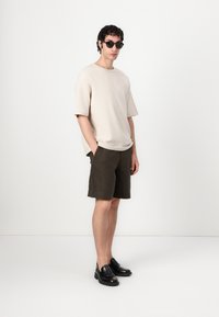Béžové tričko s krátkým rukávem v kombinaci s tmavě zelenými šortkami, obuté s černými mokasíny a slunečními brýlemi. Minimalistický design, volný střih.