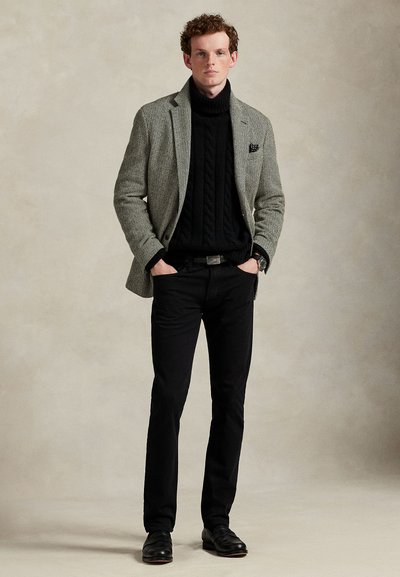 Grå fiskebeinsmønstret blazer over en svart kabelstrikket høyhalset genser, kombinert med svarte bukser og svarte sko. Merkbar klokke på håndleddet.