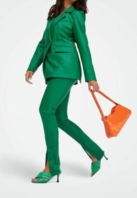 Blazer in ecopelle verde e pantaloni abbinati con fessure. Borsa arancione brillante e sandali verde con tacco a stiletto. Texture liscia, vestibilità sartoriale.