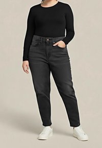 Mujer vestida con top negro de manga larga, jeans negros de cintura alta, zapatillas blancas, de pie con una mano en el bolsillo contra un fondo liso.