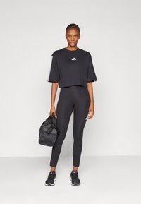 Μαύρο κοντό μπλουζάκι με λευκό λογότυπο Adidas, συνδυασμένο με μαύρες κολάν που διαθέτουν λογότυπο στη λεπτομέρεια. Το μοντέλο κρατά μια μαύρη αθλητική τσάντα και φορά αθλητικά παπούτσια.