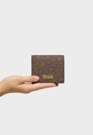 Ręka trzymająca brązowy, teksturowany, kwadratowy portfel z złotym logo "TOUS" umieszczonym na środku przedniej części, na jednolitym jasnym tle.