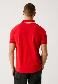 Crvena polo majica s čvrstom teksturom, sa uskim ovratnikom i bijelim obrubom, prikazuje logo "PEPE JEANS" na stražnjem dijelu ovratnika.