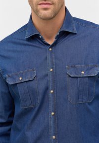 Camicia in denim blu scuro con colletto strutturato, due tasche sul petto e bottoni metallici. Presenta cuciture a contrasto.