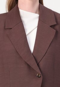 Hnědý blazer s velkými klopami, zapínání na jeden knoflík a texturovaná tkanina. Nosený přes krémový vršek, vyzdvihující vrstvený vzhled.