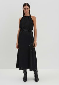 Abito nero con scollo a halter dallo stile morbido, stretto in vita con una cintura sottile. Il tessuto appare liscio e leggero. Lunghezza fino al ginocchio.