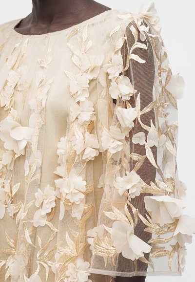 Διαφανές μπεζ ύφασμα με τρισδιάστατες λουλουδένιες εφαρμογές και φύλλα με χρυσή κεντητική, draped πάνω σε σκούρο δέρμα, καλύπτοντας τον ώμο και το άνω χέρι.