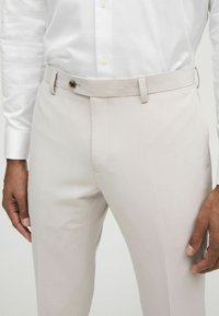 Pantalones de color beige claro con corte ajustado, frente plano y cierre de botón en la cintura. Tela suave y detalle de costura sutil visible.