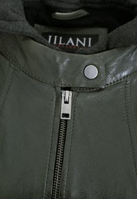 Olivgrüne Lederjacke mit hohem Kragen, versehen mit einem silbernen Reißverschluss und Druckknopfdetails. Das Innere ist mit weichem grauem Stoff ausgekleidet.