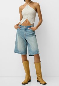 Jasny beżowy top na szyję z dzianiny, jasnoniebieskie szorty jeansowe z wysokim stanem oraz beżowe kozaki do kolan na słupku.