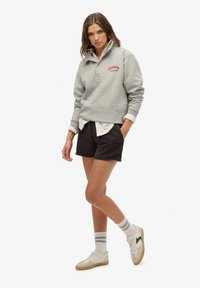 Szara bluza z wysokim kołnierzem i czerwonym logo "Superdry", połączona z ciemnymi szortami i białymi skarpetkami w zielone paski, noszona z trampkami.
