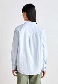 Lys blå, langermet skjorte med bakfold, knappelommer og buet kant. Laget av en glatt bomullsblanding. Enkel og ren design.