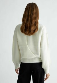 Maglione bianco a coste con design a V sul retro, maniche lunghe a sbuffo e orlo aderente; indossato con pantaloni neri, per un look casual.