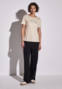 Camiseta de algodón beige con el texto "Colores de Temporada", combinada con pantalones negros holgados. El conjunto se completa con zapatos casuales de color claro.