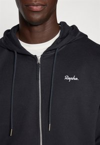 Czarna bluza zip-up wykonana z miękkiego materiału, z białym haftowanym logo i regulowanymi sznurkami. Gładka tekstura, casualowy design.