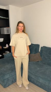 Beige T-shirt met de tekst "PECADOR", gecombineerd met tan geruit wijde broekspijpen en witte sneakers, op een teal corduroy bank.