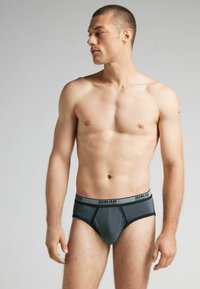 Grijze katoenen slip met zwarte rand en logobund. Heeft een nauwsluitende pasvorm en naadloos ontwerp. Model poseert zijwaarts en toont het zijprofiel.