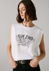 Donna con capelli corti castani, che indossa una maglietta senza maniche bianca con una grafica "Pepe Jeans London" e pantaloni neri con una cintura, mani nelle tasche.