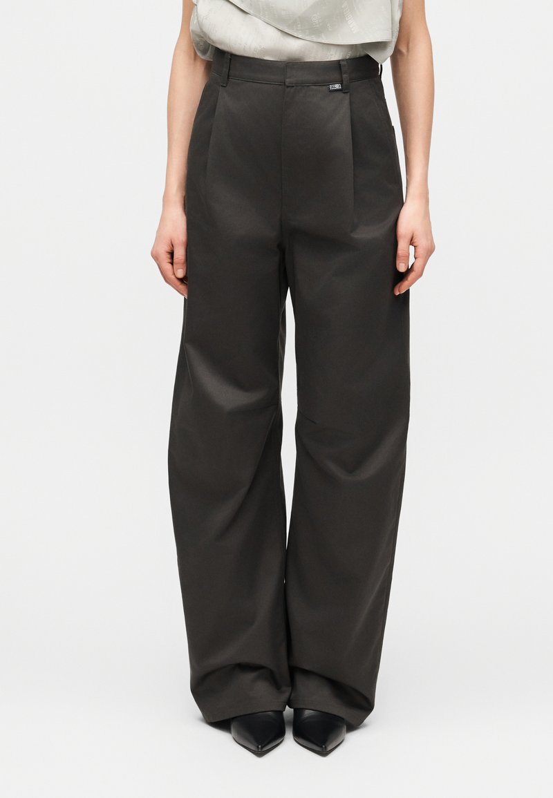 Calças de cintura alta e pernas largas em tecido cinza escuro. Apresentam pregas frontais, bolsos laterais e uma pequena etiqueta com a marca na cintura.