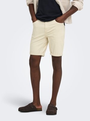 Homme portant un short beige à ourlet roulé, une chemise noire, une veste légère et des chaussures marron foncé sans lacets, debout les mains dans les poches.