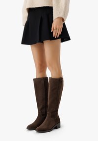 Brązowe zamszowe botki do kolan na słupku, z bocznym zamkiem błyskawicznym i przeszyciami, zestawione z czarną spódnicą plisse.