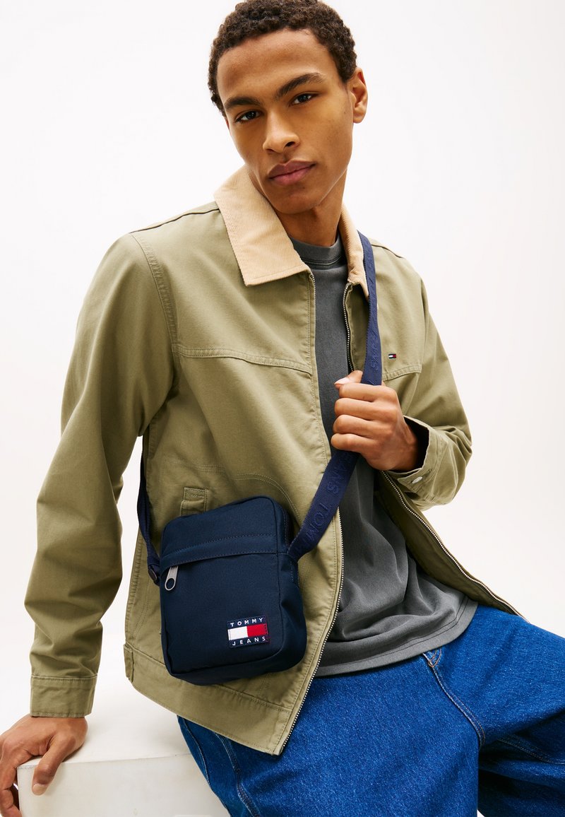 Jonge man met een olijfgroene jas en blauwe spijkerbroek met een marineblauwe Tommy Jeans crossbodytas, zittend tegen een witte achtergrond.