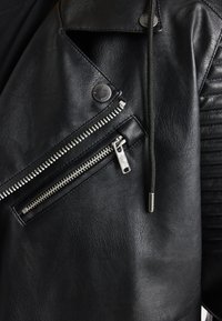 Giacca da motociclista in pelle nera con tasca con zip diagonale, dettagli testurizzati sulle spalle, accessori in argento e una leggera texture granulosa.