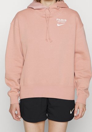 Persona vestida con una sudadera rosa del Paris Saint-Germain y pantalones cortos negros de Nike, de pie con los brazos relajados a los lados.