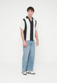 Κοντομάνικο πόλο πουκάμισο με κάθετες ρίγες σε ναυτικό μπλε και λευκό, V-λαιμόκοψη και κόκκινο κεντημένο λογότυπο. Συνδυάζεται με φαρδιά ξεβαμμένα τζιν παντελόνια και αθλητικά παπούτσια.