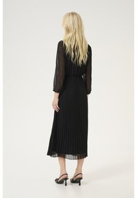 Schwarzes, plissiertes Midikleid mit transparenten langen Ärmeln, tailliertem Bund und schwingendem Rock. Getragen mit schwarzen Absatzsandalen.