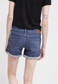 Person som bär uppkavlade jeansshorts och en svart tröja, visar en liten tatuering av planeter och stjärnor på överarmen.