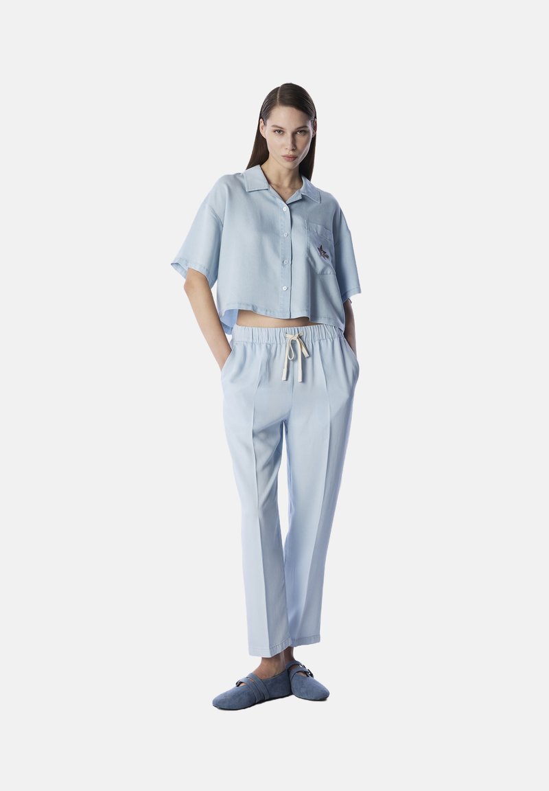 Camicia button-up azzurro chiaro, corta con maniche corte e tasca applicata, abbinata a pantaloni larghi coordinati e morbidi pantofole blu.