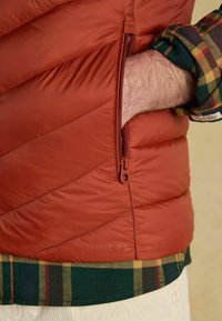 Prešívaná pufová bunda v hrdzavej oranžovej farbe s postranným zipsom a logom. Spárovaná so zelenou károvanou flanelovou košeľou, ktorá ukazuje manžety s kontrastnými farbami.