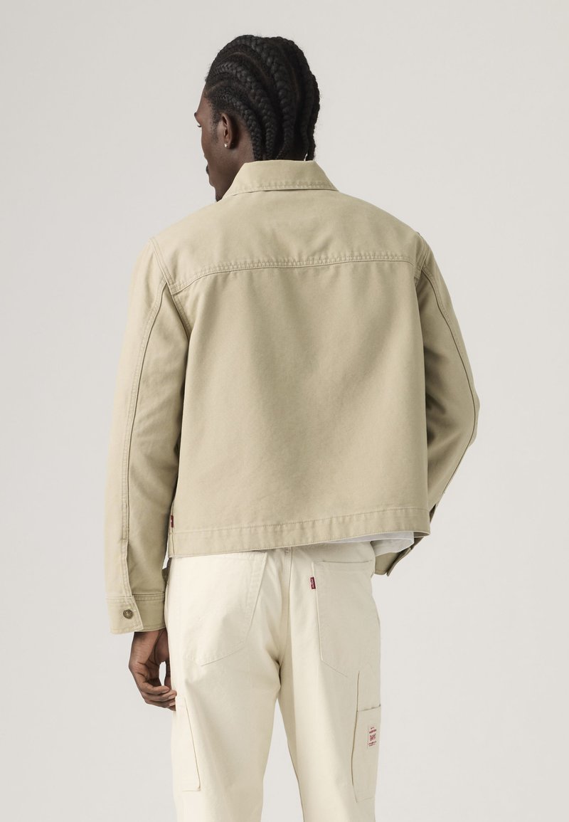 Mann mit geflochtenem Haar, der eine beige Jacke und off-white Hosen trägt, von hinten vor einem einfarbigen hellen Hintergrund gesehen.