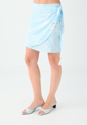 Femme portant une jupe portefeuille à motif bleu clair et blanc avec un lien à pompons et des sandales bleu clair à talons moyens et à bout ouvert, debout sur un fond blanc.