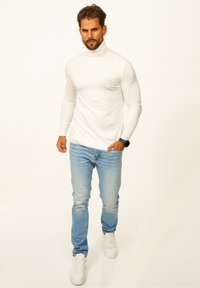 Weißer figurbetonter Rollkragenpullover, hellblaue Jeans und weiße Sneaker. Das Outfit wird durch eine schwarze Armbanduhr abgerundet.