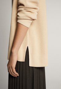 Pull beige en tricot avec poignets et ourlet côtelés. Dispose d'une fente latérale et d'un tissu texturé. Porté sur une jupe plissée foncée.