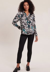 Overhemd met knopen en een zwart-wit bloemenpatroon met turquoise accenten. Gecombineerd met strakke zwarte broek en zwarte instappers.