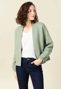 Cardigan trapuntato verde chiaro con scollo rotondo e apertura frontale, morbido al tatto e con polsini elasticizzati, abbinato a una maglietta bianca a scollo a V e jeans scuri.