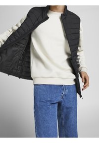 Svart puffer-väst med quiltat mönster, olåst över en krämfärgad sweatshirt, ihop med blå denimjeans. En avslappnad, lager-på-lager-outfit.