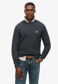 Marineblauw sweatshirt met ronde hals en geribde manchetten, laag over een licht overhemd met kraag. Bevat een klein logo-op-patch op de borst.