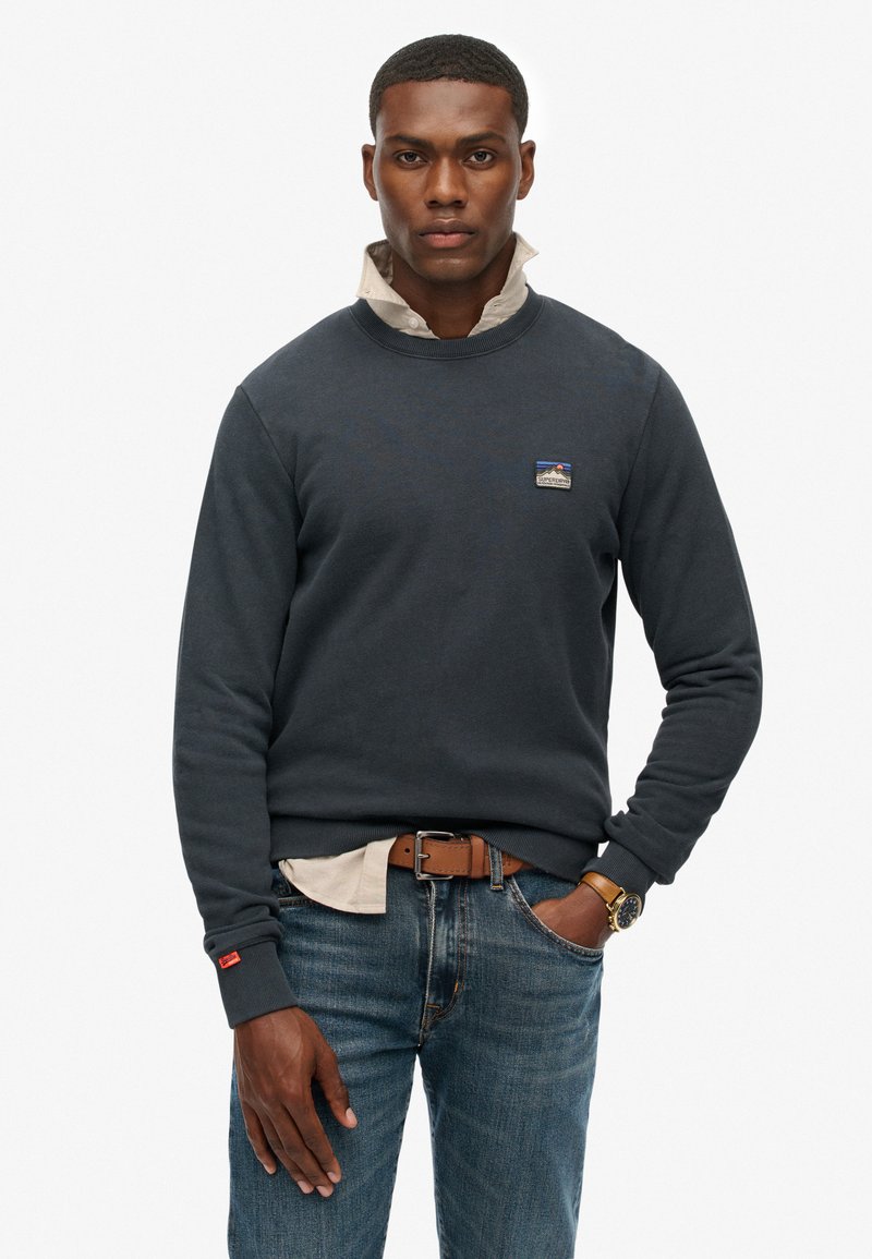 Marineblauw sweatshirt met ronde hals en geribde manchetten, laag over een licht overhemd met kraag. Bevat een klein logo-op-patch op de borst.