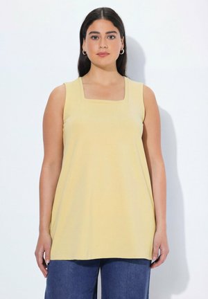 Frau mit langen dunklen Haaren, die eine ärmellose gelbe Tunika mit quadratischem Ausschnitt und blaue Jeans trägt, steht vor einem weißen Hintergrund.