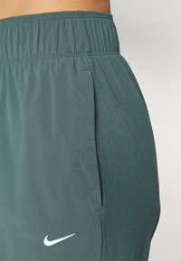Nike sporta bikses tumši zaļā audumā. Aprīkotas ar elastīgu jostu, sānu kabatām un baltu logo kreisajā pusē. Gluda tekstūra.