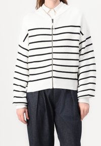 Biały kardigan robiony na druty z czarnymi poziomymi paskami, z zamkiem z przodu, ściągaczami na rękawach i skróconym fasonem. Połączony z luźnymi ciemnymi spodniami.