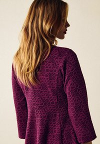 Luumuinen mekko, jossa on pitkät hihat ja kohokuvioitu kuvio korotetuista kukista ja pisteistä, valmistettu pehmeästä materiaalista, kuvattu vinosti takaa.