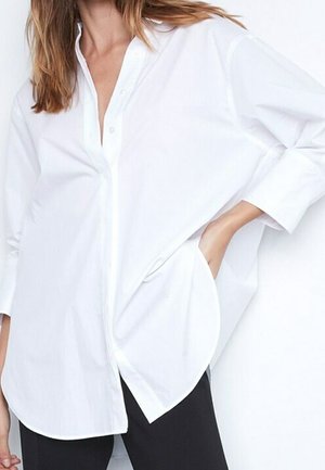Woman wearing oversized white button-up shirt with long sleeves, one hand in pocket, paired with black pants.