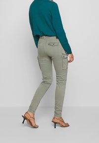 Khaki cargobroek met zijzakken, aansluitend ontwerp en een gladde textuur, gecombineerd met een smaragdgroene trui en sandalen met hoge hakken.