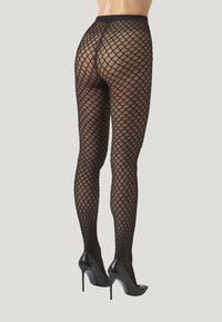 Leggings noirs en résille présentant un motif écailleux, à taille haute et avec une texture transparente, associés à des chaussures à talons hauts noires.